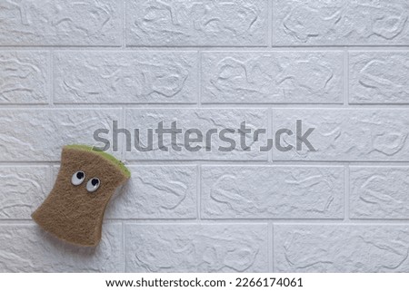 Washcloth for cleaning with cheerful face. Concept fun cleaning. Copy space. Cartoon kitchen sponge with eyes on white brick wall background. Flat lay. Template for advertising, blog, article, cover.