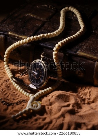 Time to pray. Pocket watch with prayer beads and book on sands.
