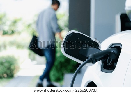 Focus electric car charging at home charging station with blurred progressive man walking in the background. Electric car using renewable clean for eco-friendly concept. Royalty-Free Stock Photo #2266203737