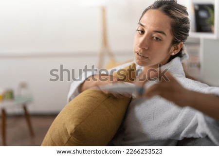 Young lady wearing a white robe, very ill, looking at the thermometer, after taking her temperature, sitting in the living room of her apartment.