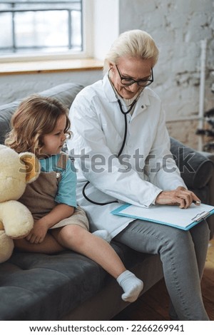 Kind female senior paediatrician doctor visiting his patient at home, examining little girl, writing prescription. Concept of kid's health check. Measure temperature, heart beat Royalty-Free Stock Photo #2266269941