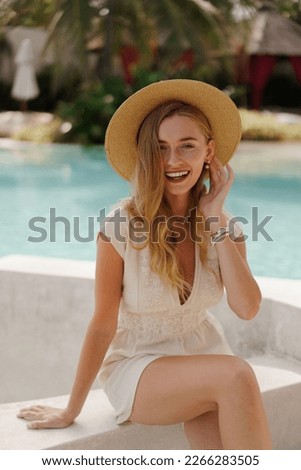 Pretty smiling blond  traveling woman in straw hat posing  near swimming pool . White summer clothes. Perfect wavy hairs.