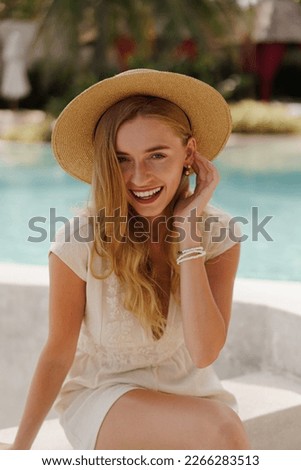 Pretty smiling blond  traveling woman in straw hat posing  near swimming pool . White summer clothes. Perfect wavy hairs.