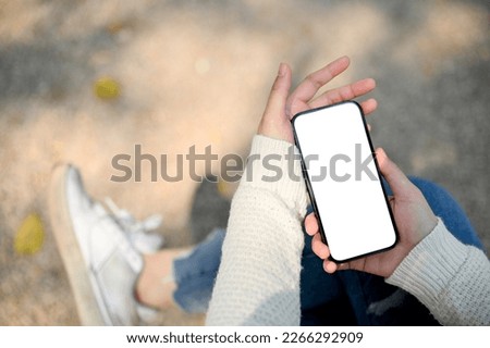 Close-up image of a woman's hand in white sweater holding a smartphone white screen mockup over blurred street in background. people and technology concept
