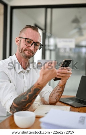 Thoughtful Caucasian man with glasses and tattoo on his arms holding a credit card or debit card and his smartphone at his desk. 