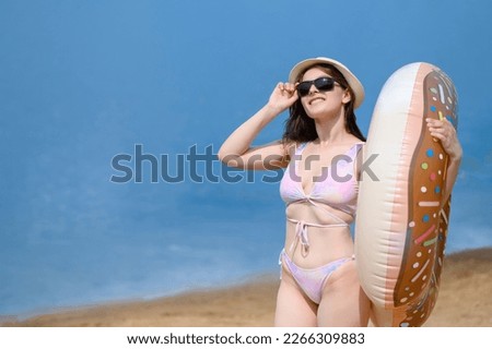 A young woman in a swimsuit, hat, sunglasses stands on the shore with an inflatable big donut near the water. The concept of vacation, rest, relaxation, resorts, water activities, weekends