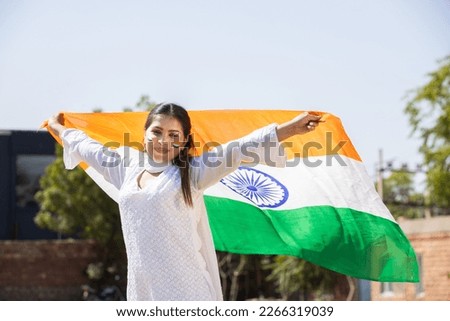 Happy young woman wearing traditional white dress weaving indian flag outdoor at park, celebrating Independence day or Republic day.