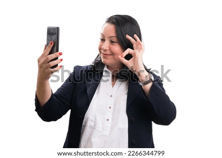 Smiling friendly businesswoman wearing casual suit during videocall using smartphone making okay gesture with fingers isolated on white background