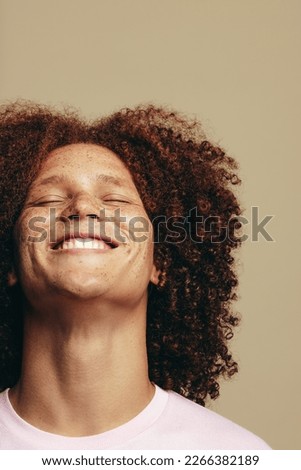 Young man with freckles and curly ginger hair smiles with his eyes closed in a studio, radiating self-love and self-confidence. Happy young man embracing his unique skin, hair and beauty features. Royalty-Free Stock Photo #2266382189