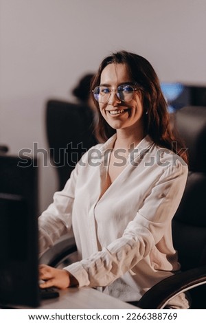 Beautiful secretary girl working with computer. Smiling graphic designer in glasses posing at her workplace. Indoor shot of female manager in white blouse