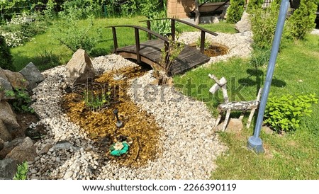 On the grassy lawn with bushes, there is a small decorative pond with water lined with rubble and stones. A wooden bridge with handrails was built over it. Sunny Royalty-Free Stock Photo #2266390119