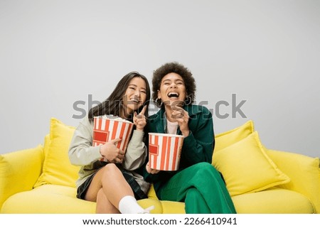 cheerful asian woman with popcorn pointing with finger while watching movie near african american friend isolated on grey Royalty-Free Stock Photo #2266410941