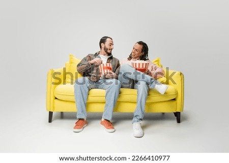full length of stylish multiethnic men smiling at each other while sitting with buckets of popcorn on yellow couch on grey background Royalty-Free Stock Photo #2266410977