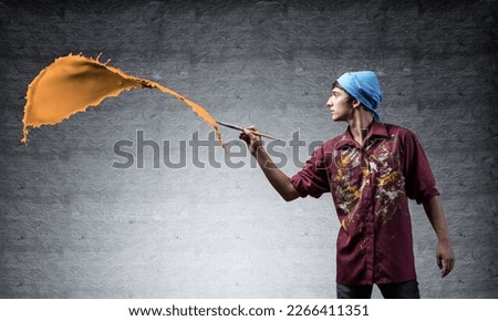 Young artist painting orange brush stroke. Male painter in dirty shirt and bandana with paintbrush on grey wall background. Creative hobby and artistic occupation. Art classes concept with copy space.