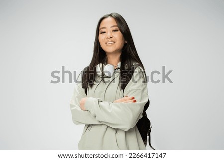 pretty asian student with wireless headphones and backpack posing with crossed arms isolated on grey Royalty-Free Stock Photo #2266417147