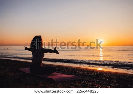 Carefree woman sitting in yoga pose with open arms by the sea sunrise time. Finding inner peace spiritual healing lifestyle. Stretching in the morning for increasing energy. Royalty-Free Stock Photo #2266448225