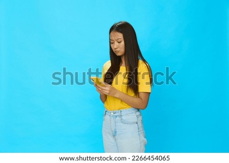 Happy Asian woman holding a phone and looking at the screen with a yellow case on a blue background in a yellow T-shirt smiling with teeth 