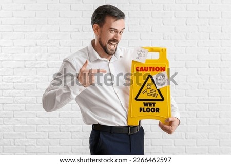 Young man pointing at caution sign with sticky papers on white brick background. April Fools' Day celebration