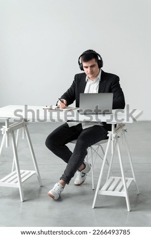 Focused young european man wear headphones, studying online, watching webinar podcast on laptop, listening to study, education course, conference calling, taking notes, sitting at desk
