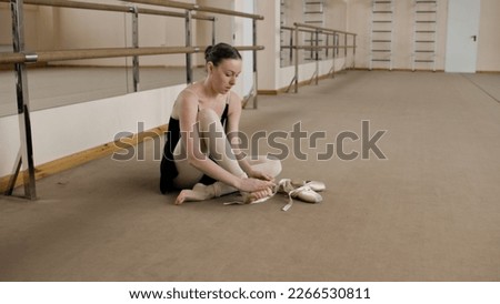 Female ballet dancer finished choreography rehearsal. Tired ballerina in training bodysuit takes off pointe shoes after stretching and gymnastic workout in dance studio. Classic ballet dance school. Royalty-Free Stock Photo #2266530811