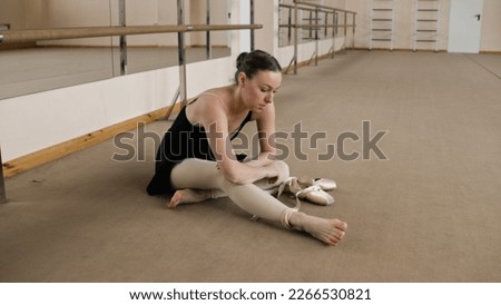 Female ballet dancer finished choreography rehearsal. Tired ballerina in training bodysuit takes off pointe shoes after stretching and gymnastic workout in dance studio. Classic ballet dance school. Royalty-Free Stock Photo #2266530821