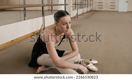 Female ballet dancer finished choreography rehearsal. Tired ballerina in training bodysuit takes off pointe shoes after stretching and gymnastic workout in dance studio. Classic ballet dance school. Royalty-Free Stock Photo #2266530823