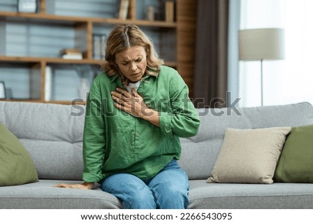 Senior beautiful woman sitting sick on sofa at home. Holds his chest. He has asthma, allergies, feels severe pain, tenderness, panic attack, infarct. Royalty-Free Stock Photo #2266543095