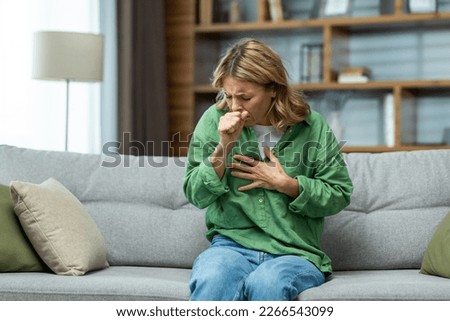 Senior beautiful woman sitting sick on sofa at home. He coughs, covers his mouth with his hand, holds his chest. Feels pain, suffers from asthma, allergies, flu, cold. Royalty-Free Stock Photo #2266543099