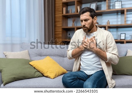 A young man is sitting on the sofa at home and holding his chest. Feels severe pain, shortness of breath, panic attack, heart attack, stroke. Royalty-Free Stock Photo #2266543101