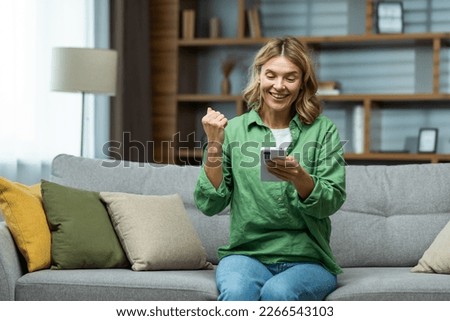 A happy senior woman is sitting on the sofa at home, holding the phone in her hands. Shows a victorious yes gesture with his hand.