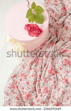  A round delicate pink box and a rose flower next to dresses in pink flowers. Focus on the box.