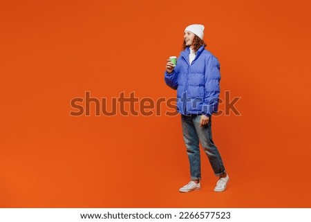 Full body side view young man with long curly hair wear hat purple ski padded jacket casual clothes hold takeaway delivery craft paper brown cup coffee to go isolated on plain orange red background