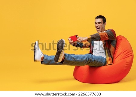 Full body young middle eastern man wear casual shirt t-shirt using play racing app on mobile cell phone hold gadget smartphone for pc video games sit in bag chair isolated on plain yellow background