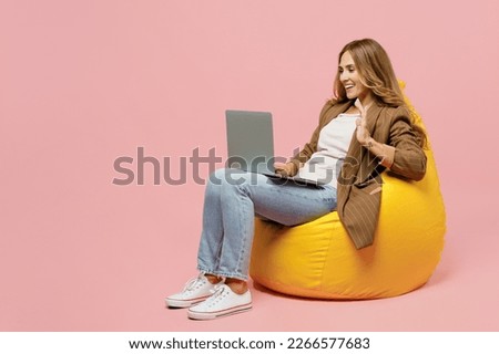 Full body young employee business woman she wear casual brown classic jacket sit in bag chair hold use work on laptop pc computer talk video call waving hand isolated on plain pastel pink background