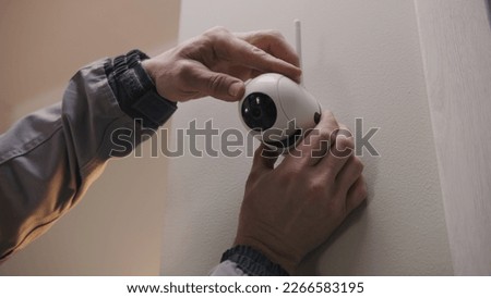 Installer in uniform puts security camera on wall fastening and connects it to system with cable. Man installs cameras in house. Concept of CCTV cameras, monitoring, safety and privacy. Royalty-Free Stock Photo #2266583195