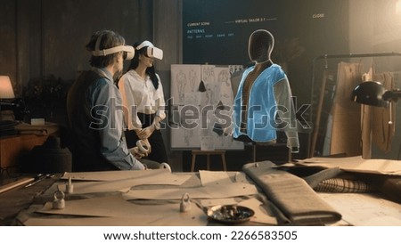 Tailor and female customer put VR goggles in atelier workshop. 3D hologram of jacket pattern. Virtual interface menu of program for designing and modeling clothes. Technologies of augmented reality. Royalty-Free Stock Photo #2266583505