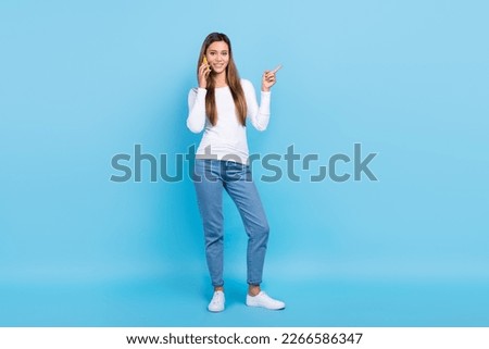 Full length photo of shiny cute woman wear white shirt talking gadget pointing empty space isolated blue color background