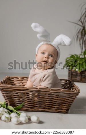 baby in a white bodysuit with rabbit ears on his head is sitting in a wicker basket on the floor with a bouquet of tulips. High quality photo