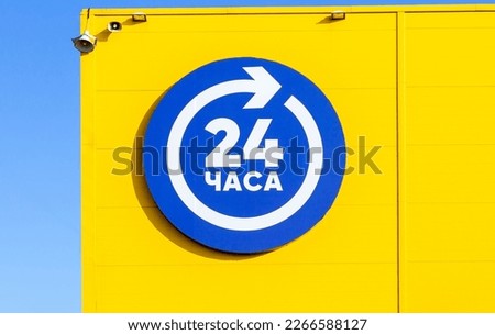 24 hours blue sign open or working around the clock hanging on yellow wall. Shop working 24 hours, sign on facade of building. Inscription on russian: 24 hours