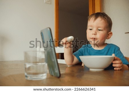 Child, a boy of three years old, is having breakfast at the kitchen table and watching cartoons on his smartphone