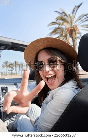 Vertical portrait pretty young hipster big smile in convertible car. Beautiful woman with sunglasses and hat making peace sign. People renting car for vacation on sunny day. Funny moments outdoors.