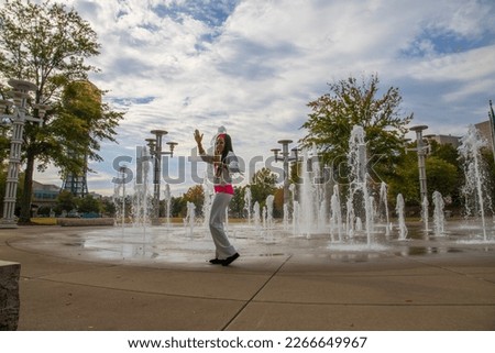 an African American woman with long sisterlocks wearing white and pink clothes, sunglasses and an orange head scarf holding dancing in front of a water fountain with lush green trees	