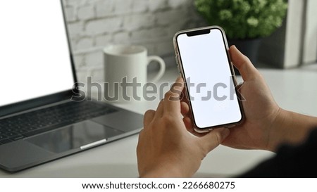 Close up view of hands holding smart phone with white empty screen in front of laptop computer on white table