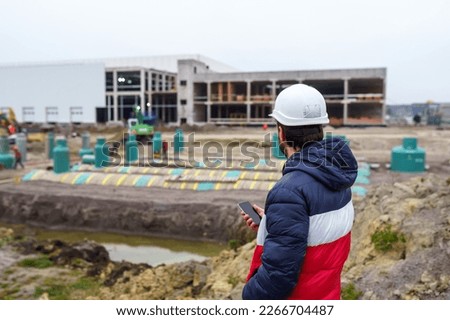 Engineer builder in a hard hat with a smartphone takes a photo on the background of the construction of an industrial building