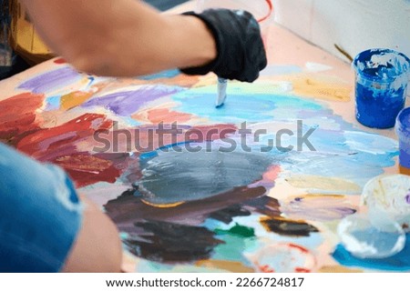 Artist hand in black gloves holds paint brush and choose color from colorful palette at outdoor art painting festival, paintings art picture process. Artist paints atmospheric surreal picture