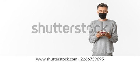 Concept of covid-19, quarantine and holidays. Image of dreamy middle-aged man in medical mask and glasses, looking amazed at birthday cake, making a wish, standing over white background.