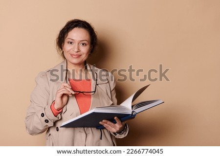 Confident positive middle-aged multi-ethnic female teacher, professor, educator, holding eyeglasses and book with fairy tails, smiling looking at camera, isolated over beige color background
