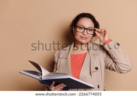 Multi-ethnic beautiful middle-aged woman, a teacher, professor, educator, holding a hardcover book with fairy tails, and smiling looking at camera through eyeglasses, isolated beige color background