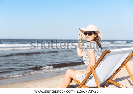 Happy brunette woman wearing sunglasses while relaxing on a wooden deck chair at the ocean beach.