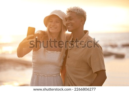 Closeup portrait of an young affectionate mixed race couple standing on the beach and smiling and taking a selfie with a smartphone during sunset outdoors. Hispanic man and caucasian woman showing l